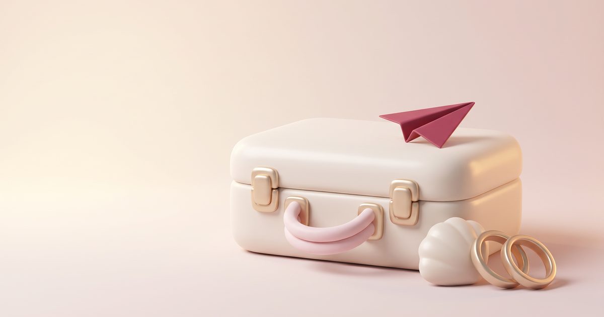 A cream clay suitcase with gold clasps, a rose paper airplane on its lid, a seashell and two gold rings