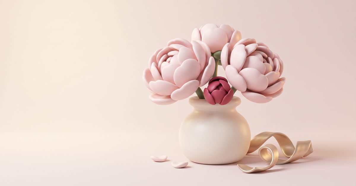 A cream vase of blush peonies and a rose bud beside a loose gold ribbon and two fallen petals